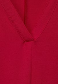 Top con scollatura a V di un vivace colore rosso. Realizzato in tessuto morbido e liscio con cuciture visibili lungo il collo e una sottile texture.
