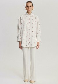 Camicia bianca con ricami floreali rossi, vestibilità comoda e maniche lunghe. Abbinata a pantaloni a campana crema e scarpe beige.