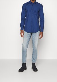 Camicia button-up blu navy a maniche lunghe con colletto, abbinata a jeans dritti blu chiaro e stivali neri alla caviglia.