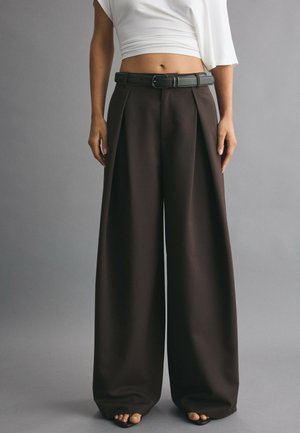Person iført højtsiddende, vidde mørkebrune bukser med sort bælte, en hvid cropped top og åbne sko på en grå baggrund.