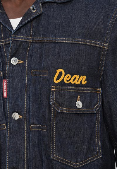 Denimjakke med mørkeblåt stof, gul syning og broderet "Dean" på brystlommen. Sølv snapknapper er til stede.