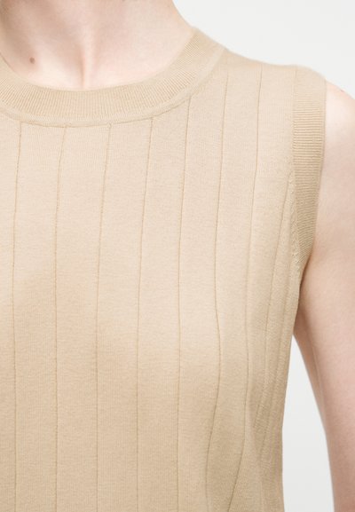 Κοντινό πλάνο ενός ατόμου που φοράει μπεζ αμάνικο ριμπ πλεκτό τοπ με στρογγυλή λαιμόκοψη πάνω σε απλό φωτεινό φόντο.