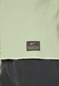 Haut à manches longues en tricot gaufré vert clair avec une étiquette noire portant un texte jaune au bas, associé à un pantalon gris foncé.