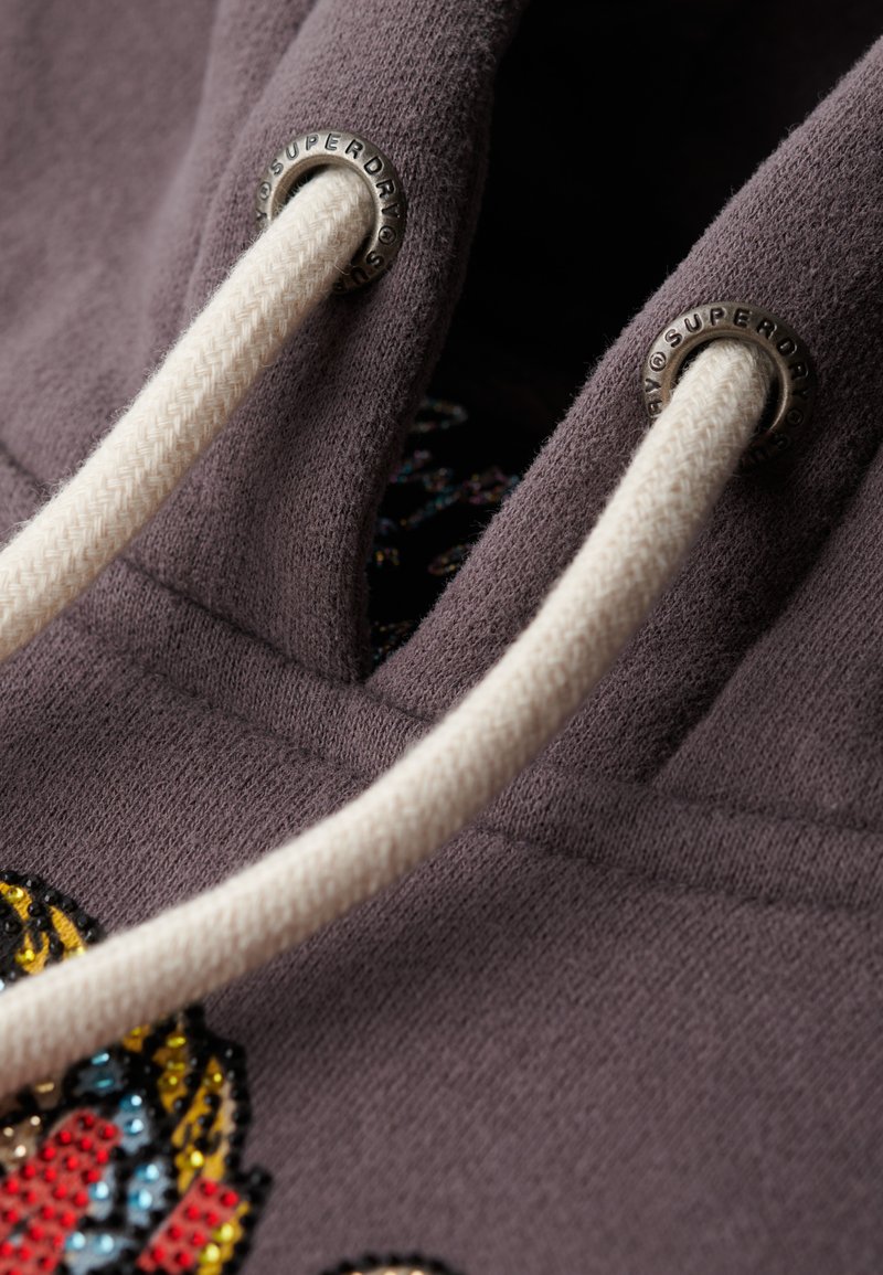 Zbliżenie ciemnofioletowego hoodie z białymi sznurkami przewleczonymi przez metalowe oczka z wygrawerowanym napisem "Superdry", ukazujące część kolorowego wzoru.