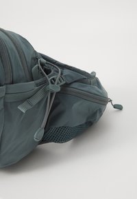 Zaino turchese con un esterno in nylon liscio. Presenta una tasca in rete e spallacci regolabili. Include dettagli con zip e una forma slanciata.