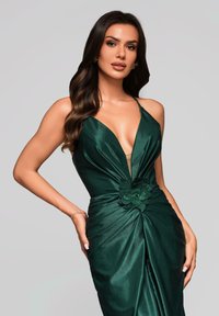 Vrouw met lang donker haar draagt een mouwloze diep groene satijnen jurk met een diep decolleté en bloemdetaillering bij de taille, poseert tegen een grijze achtergrond.