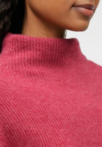 Adīta džemperis dziļi rozā krāsā ar augstu apkakli un smalku ribu tekstūru, demonstrējot gludu un mīkstu audumu.