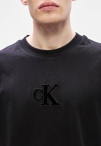 Melna kokvilnas T-krekls ar apaļu kakla izgriezumu, kurā ir iespiests melns "cK" logotips uz krūšu, gluda tekstūra un īsas piedurknes.