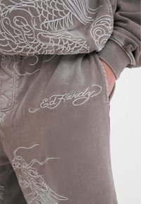 Graue Sweatpants mit aufwändiger weißer Stickerei eines Drachen und anderen Mustern, mit einem sichtbaren Logo, das "Ed Hardy" zeigt. Weicher Stoff.