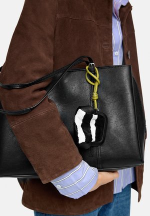 Osoba ubrana w brązową zamszową kurtkę i niebieską koszulę w paski trzyma czarną skórzaną torbę typu tote z breloczkiem w czarno-białe wzory.