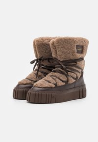 Braune Stiefel mit Futter aus Fell, knöchelhochelend, mit einer strukturierten, klobigen Sohle, die dunkle Schnürsenkel und ein Markenpatch an der Seite aufweisen.