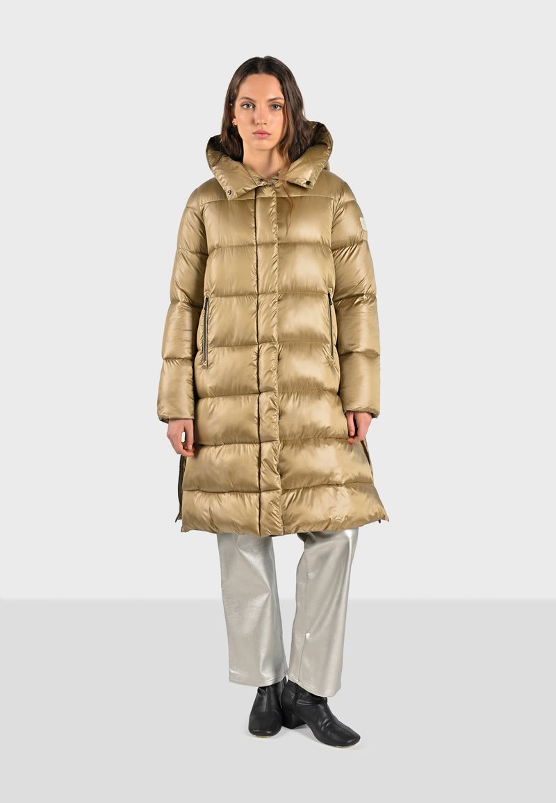 Manteau matelassé brun clair avec capuche, design matelassé, fermeture éclair devant et poches latérales. Associé à un pantalon argenté et des bottines noires.