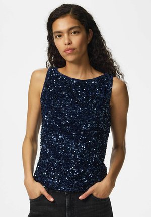 Femme aux longs cheveux bouclés portant un haut sans manches bleu foncé à sequins, les mains dans les poches d’un jean noir, face à l’avant.