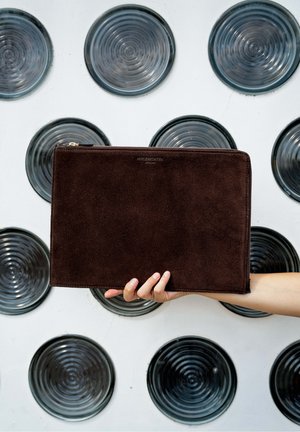 Hand holding a dark brown rectangular suede pouch with zipper, against a white wall with black circular patterns.