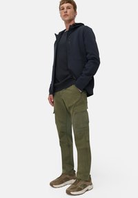 Marineblaue Kapuzenjacke mit einer Reißverschlusstasche, über einem dunklen Oberteil getragen, kombiniert mit olivgrünen Cargo-Hosen und braunen Sportschuhen mit Akzenten.