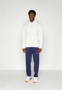 Homem de pé contra um fundo branco, vestindo um hoodie branco, calças de treino azul-marinho e ténis brancos com solas de borracha.