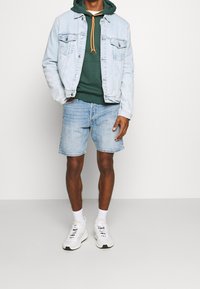 Giacca di jeans azzurro chiaro con felpa con cappuccio arancione e coulisse, pantaloncini di jeans azzurro chiaro e sneakers bianche con dettagli neri.