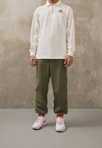 Polo de manga larga de Nike en blanco con cuello con cremallera; pantalones holgados verde oliva; zapatillas blancas con detalles rojos y cordones.