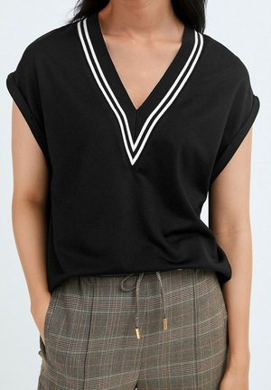 Haut noir à manches courtes avec double rayures blanches sur col en V, porté avec un pantalon à carreaux marron taille haute avec cordon et mains dans les poches.
