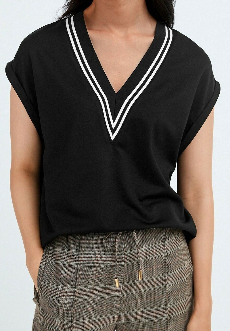 Haut noir à manches courtes avec double rayures blanches sur col en V, porté avec un pantalon à carreaux marron taille haute avec cordon et mains dans les poches.