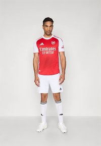 Mann in einem roten und weißen Arsenal-Fußballtrikot mit Emirates-Sponsor, weißen Shorts, Socken und Fußballschuhen, der vor einem schlichten Hintergrund steht.