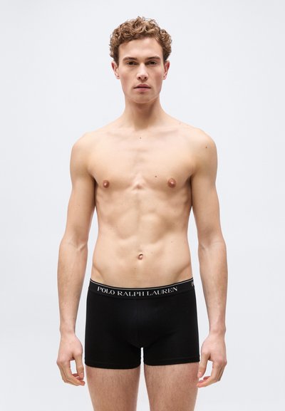 Νέος άνδρας με σγουρά μαλλιά, ντυμένος με μαύρα μποξέρ Ralph Lauren Polo, στέκεται μπροστά σε ένα απλό φωτεινό φόντο, αντιμετωπίζοντας τη κάμερα.