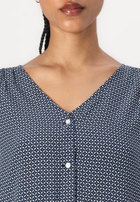 Marineblaue Bluse mit V-Ausschnitt, die ein geometrisches Muster in Hellblau und Weiß aufweist. Mit einem Knopfverschluss vorne. Leichter Stoff.
