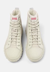 Vaalean kermaiset korkeavartiset tennarit, joissa on teksturoitu kumipohja, litteät nauhat ja punainen "CAMPER"-brändäys kielekkeessä. Sileä nahkapinta.