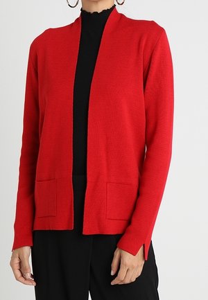 Cárdigan rojo de apertura frontal hecho de tejido de punto texturizado, con dos bolsillos frontales y aberturas laterales, llevado sobre una blusa negra.
