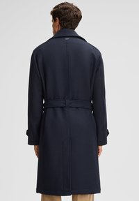 Man met de rug naar de camera, gekleed in een donkerblauwe lange wollen jas met een riem en beige broeken, tegen een effen lichte achtergrond.