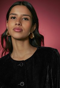 Black velvet top featuring a round neckline, two ornamental buttons, and large gold-tone triangular earrings against a pink background.