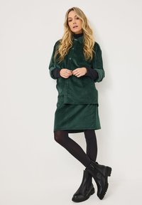 Camisa chaqueta de pana verde sobre un cuello de tortuga negro, combinada con una falda a juego y botas de tobillo negras con suelas resistentes.