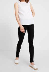 Vit ärmlös topp med spetsdetaljer, kombinerad med svarta tighta jeans och vita espadrillos. Enkla bakgrund.