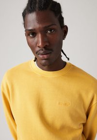 Yellow crewneck sweatshirt made of cotton, featuring a textured finish and embossed logo on the chest, worn with a white tee underneath.
