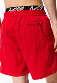 Pantaloni da bagno rossi con una cintura nera elastica che presenta un logo bianco. Include una tasca posteriore con chiusura a zip. Tessuto strutturato.