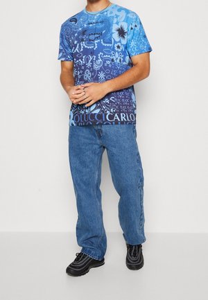 Mężczyzna w niebieskiej koszuli z krótkim rękawem w wzory, luźnych niebieskich dżinsach, czarnych trampkach i srebrnym łańcuszku, stojący z dłońmi skrzyżowanymi.