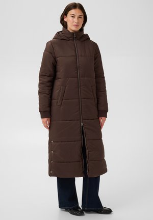Femme portant un long manteau matelassé marron avec capuche, fermeture éclair à l'avant, boutons-pression latéraux, pantalon large bleu foncé et chaussures noires.