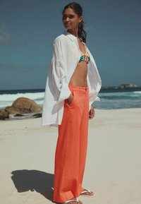 Camicia bianca oversize, bikini con motivo colorato e pantaloni larghi arancione brillante. Spiaggia sabbiosa sullo sfondo con onde oceaniche.