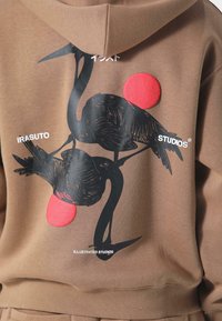 Brun hoodie med et grafisk design af to sorte fugle med røde detaljer. Tekst der læser "IRASUTO STUDIOS" og "ILLUSTRATED STUDIOS" under designet.