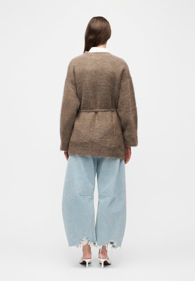 Γυναίκα με καφέ αφράτη ζακέτα με ζώνη, ανοιχτό μπλε φαρδιά τζιν παντελόνια με φθορές και λευκές μπαλαρίνες με τακούνι, στέκεται μπροστά σε λευκό φόντο.