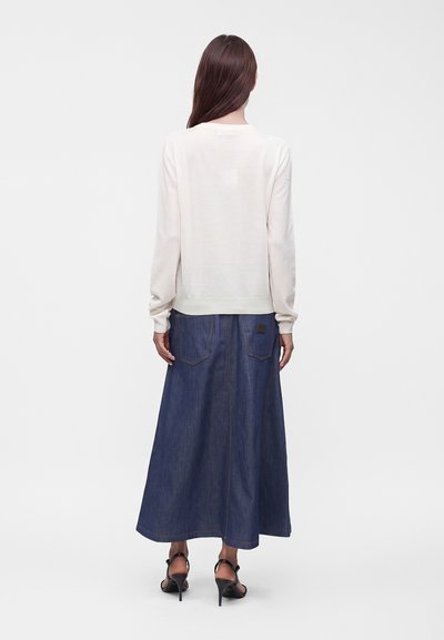 Femme aux longs cheveux bruns portant un haut blanc à manches longues, une jupe longue en denim bleu et des sandales à talons hauts noires, de dos.