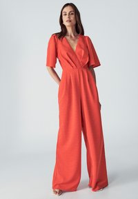 Oranje jumpsuit met korte mouwen, diepe V-hals, getailleerde taille en wijde broekspijpen; gestructureerde stof en ontspannen silhouet.