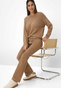 Kvinde sidder på siden på en beige stol med metalramme, iført en lys brun sweater, matchende bukser og hvide sneakers med gummisåler.