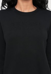 Černá mikina s žebrovaným kulatým výstřihem. Na hrudi je texturované logo "ARMANI EXCHANGE" v tmavých kamínkách. Jednoduchý design.