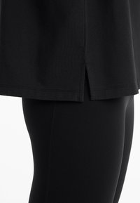 T-shirt noir avec ourlet à fente sur le côté, associé à des leggings noirs. Le tissu semble lisse et extensible, mettant en valeur une coupe ajustée.