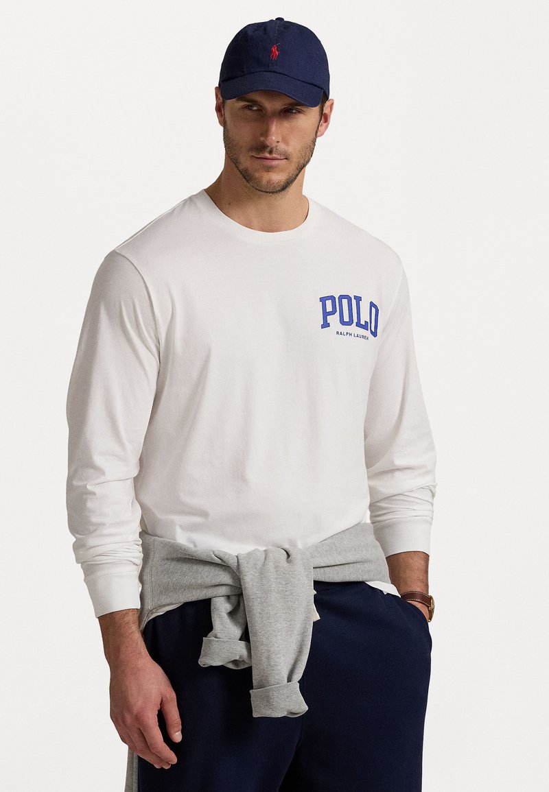 Balta ilga rankovė marškiniai su mėlynu "POLO" užrašu, pilkas megztinis surištas aplink liemenį, tamsios kelnės ir tamsiai mėlyna kepurė su raudonu logotipu. Kasdienis apranga.