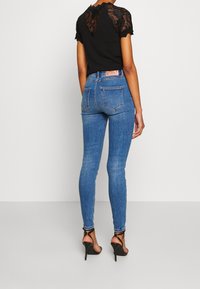 Mujer con jeans ajustados azules de tiro alto, top negro con espalda de encaje y sandalias negras de tacón alto con tiras, posando frente a un fondo liso.