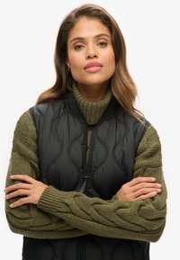 Czarny pikowany kamizelka z wysokim kołnierzem, w połączeniu z zielonym swetrem w warkocze. Kamizelka ma zamek błyskawiczny oraz strukturalne wzory faliste.