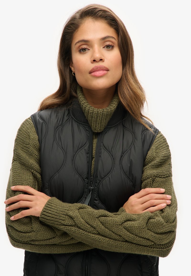 Schwarze gesteppte Weste mit hohem Kragen, kombiniert mit einem grünen Zopfmuster-Pullover. Die Weste hat einen Reißverschluss und strukturierte Wellenmuster.