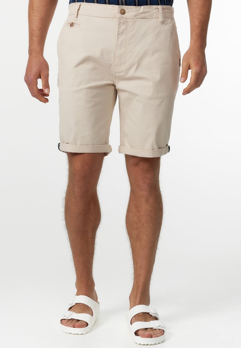 Shorts en coton beige avec des poignets roulés, des poches avant et une fermeture à bouton. Associés à des sandales blanches. Texture lisse et design décontracté.
